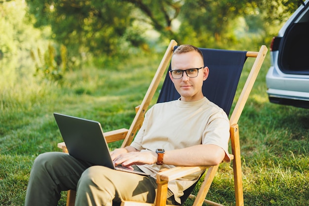 Freelancer man sits on a wooden chair in nature and works online on a laptop A man travels and works remotely on a laptop computer Office work in nature Vacation