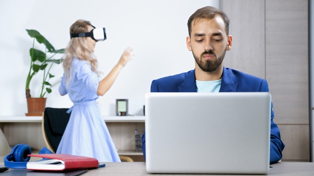 Freelancer man working at home on the computer while his wife is playing video games on the virtual reality headset