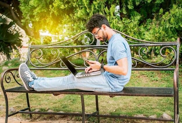 Freelancer man zit in een park met laptop en mobiele telefoon Man in een park werkt online met laptop Ontspannen man aan het werk met laptop buiten Jonge man in een park met laptop en mobiele telefoon
