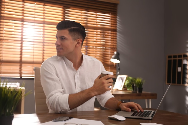 Freelancer with cup of coffee working on laptop at table indoors