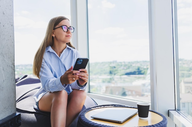 Freelancer woman in classic glasses talking on the phone and smiling while sitting in a modern coworking space carefree millennial woman in glasses enjoying leisure time for communication