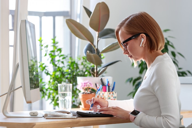 Freelancer woman taking notes with a pen, working on computer, using calculator to calculate the invoice, surrounded by plants. Distance job.