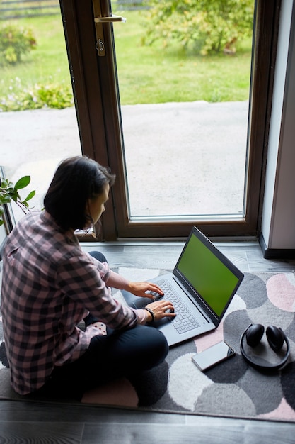 큰 창문 근처에 있는 집 바닥에 앉아 노트북에 타이핑하는 프리랜서 여성, 노트북을 검색하고, 공부하고, 집에서 일하는 콘텐츠 여성.