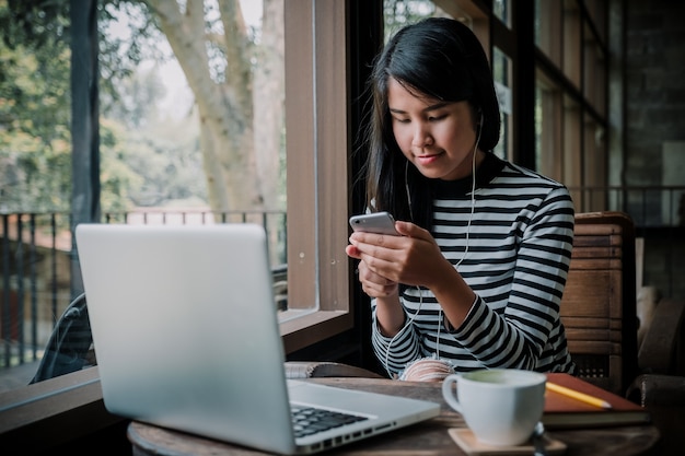 Cuffie d'uso della donna delle free lance che si siedono sul caffè che ascolta la musica su uno smartphone.