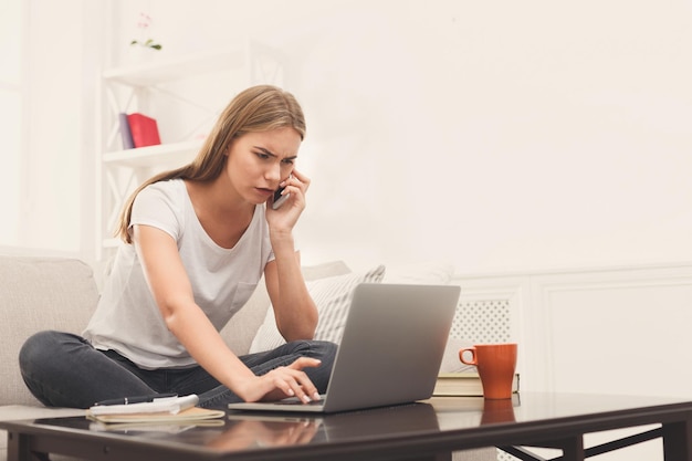 自宅でラップトップとモバイルで作業しているフリーランサー心配している金髪の女性がネットサーフィンをし、ベージュのソファのコピースペースに座って電話で話している