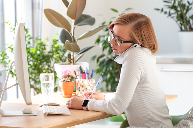 Freelancervrouw die op smartphone spreken, die nota's met een pen maken, die aan computer vanuit huisbureau werken