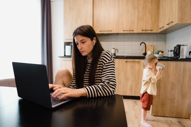 Freelancervrouw zit bij de tafel in het thuiskeukenkantoor en werkt op laptop Speels kind leidt af van het werk Kind maakt lawaai en vraagt aandacht van drukke moeder