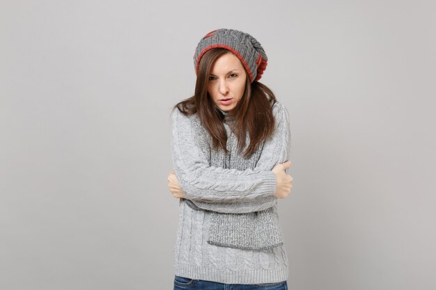 Freezing young woman in gray sweater, hat scarf holding hands folded isolated on grey background in studio. Healthy fashion lifestyle, people sincere emotions, cold season concept. Mock up copy space.