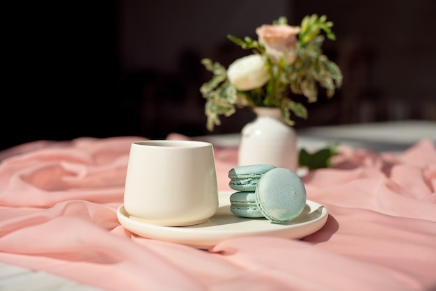 French blue macaroon and coffee cup standing on a wooden table with a pink tablecloth white vase with flowers roses and greens