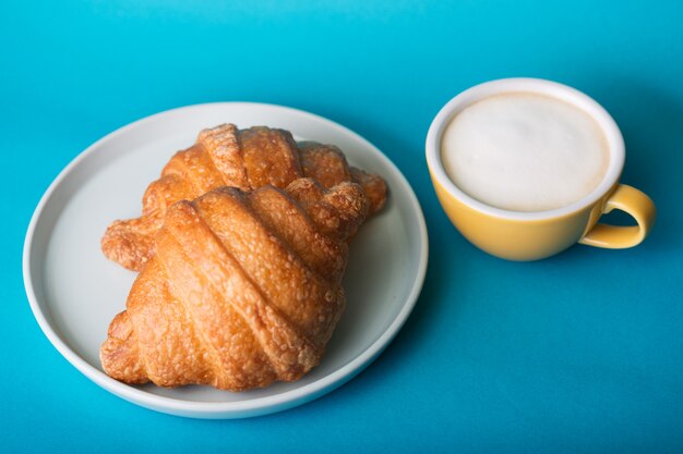 フランスの朝食。青い背景にクロワッサンと美しいコーヒーイエローカップカプチーノ