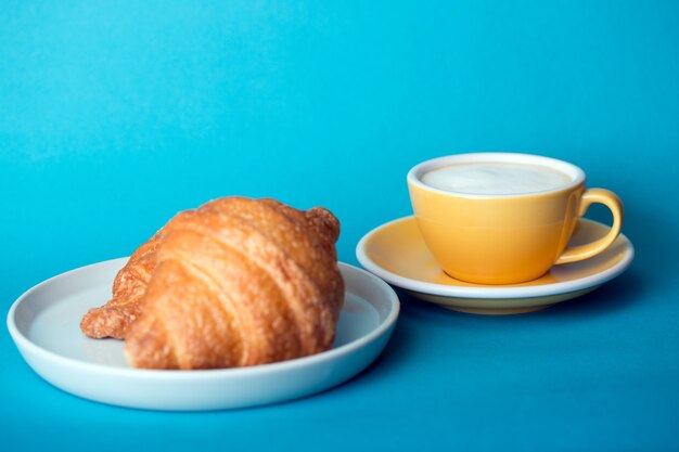 フランスの朝食。青い背景にクロワッサンと美しいコーヒーイエローカップカプチーノ