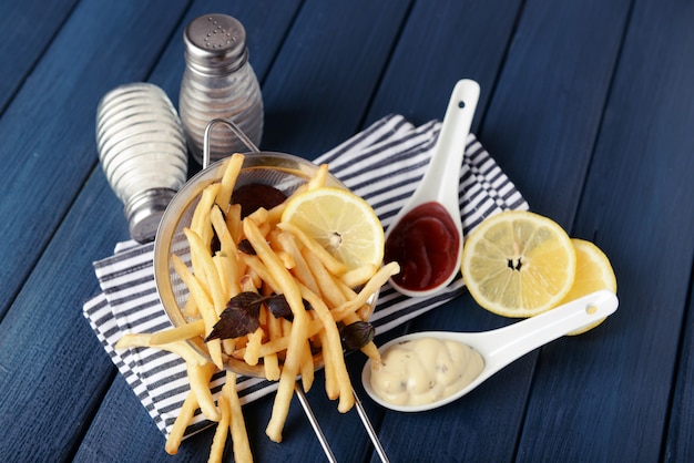 French fried potatoes in metal colander with sauce on wooden surface