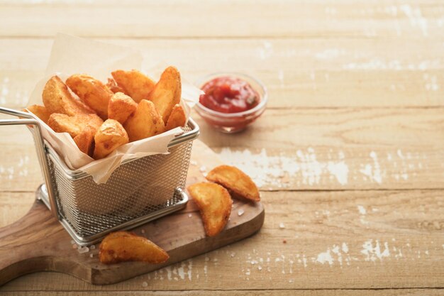 French fries in metal wire basket with salt and ketchup on old wooden light background clous up Fried potatoes Fast food and unhealthy food concept