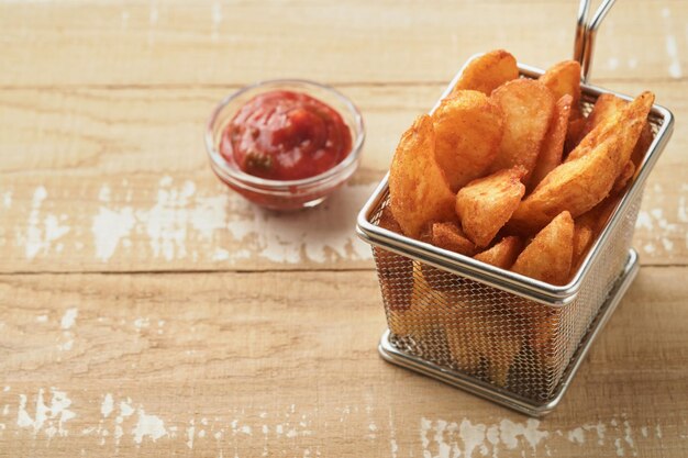 French fries in metal wire basket with salt and ketchup on old wooden light background clous up Fried potatoes Fast food and unhealthy food concept