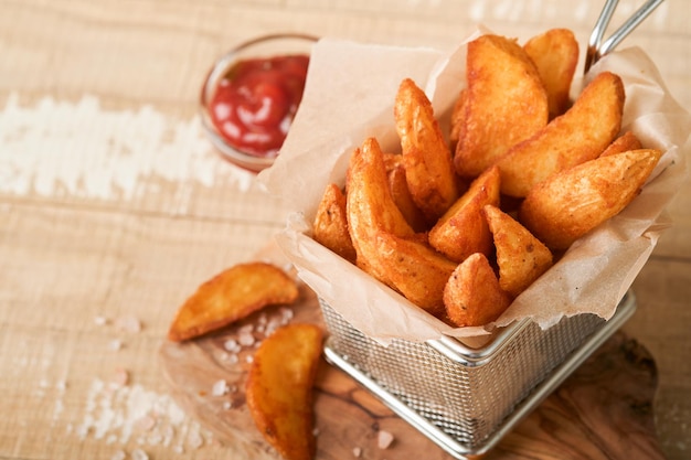 French fries in metal wire basket with salt and ketchup on old wooden light background clous up Fried potatoes Fast food and unhealthy food concept