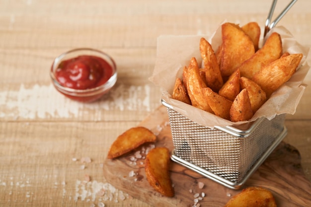 French fries in metal wire basket with salt and ketchup on old wooden light background clous up Fried potatoes Fast food and unhealthy food concept