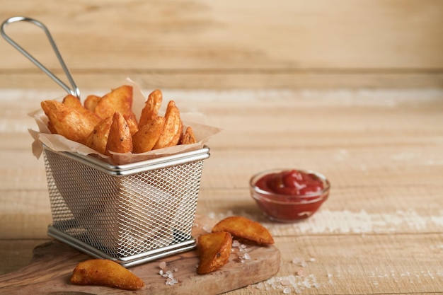 French fries in metal wire basket with salt and ketchup on old wooden light background clous up Fried potatoes Fast food and unhealthy food concept