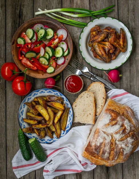 French fries, wings, salad, vegetables, bread on an old wooden background. Rural dinner, summer picnic. Top view. Flat lay.