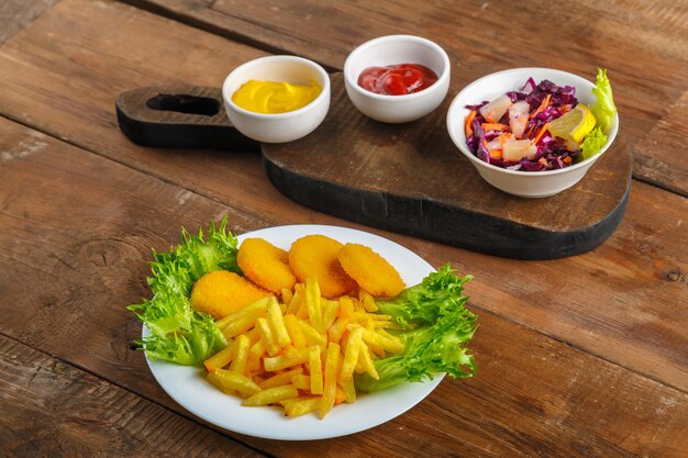 French fries with chicken nuggets next to cheese sauce and ketchup in a gravy boat and salad on wooden planks. Horizontal photo