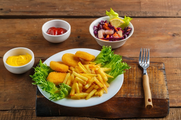 French fries with chicken nuggets next to cheese sauce and ketchup in a gravy boat and salad on a wooden table next to a fork. Horizontal photo