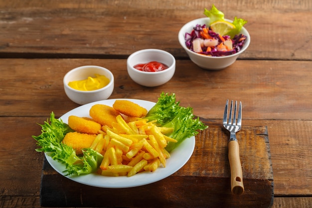 French fries with chicken nuggets next to cheese sauce and ketchup in a gravy boat and salad on a wooden table next to a fork