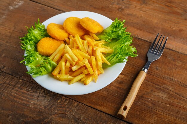 Foto patatine fritte con pepite di pollo in un piatto con insalata accanto a un tavolo di legno accanto a una forchetta. foto orizzontale