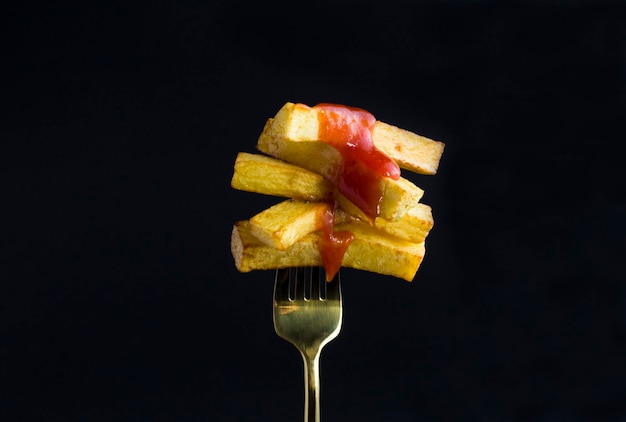 French fries with ketchup chopped on a gold fork on the black background Closeup Copy space