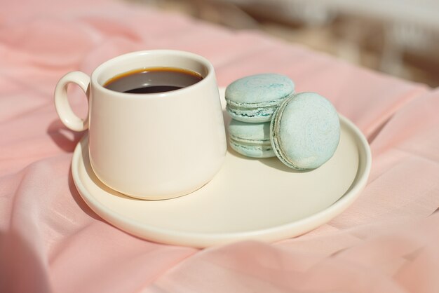 French macaroon blue plate on the pink and coffee cup standing on a wooden table with pink tablecloth white vase with flowers roses and greens.