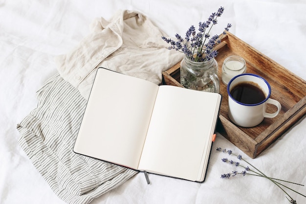 French summer still life Feminine lifestyle composition Cup of coffee lavender flowers bouquet candle on wooden tray Linen shirt nad trousers on bed Blank notebook mockup scene Top view