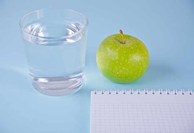 Fresh apple, glass of water and notebook on blue 