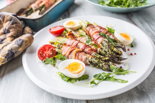 Fresh asparagus wrapped in bacon  on a white plate with arugula tomatoes and eggs.