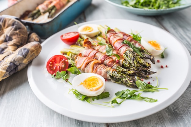 Asparagi freschi avvolti in pancetta su un piatto bianco con pomodorini e uova di rucola.