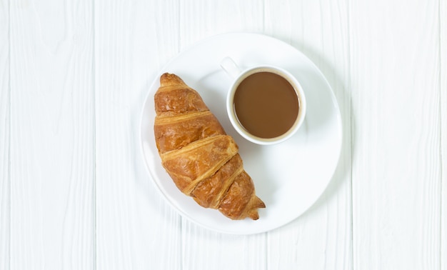 Fresh baked croissant and cup of coffee on white plate top view