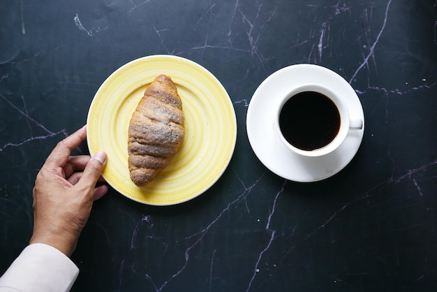 Fresh baked croissant on plate and cup of tea on black background