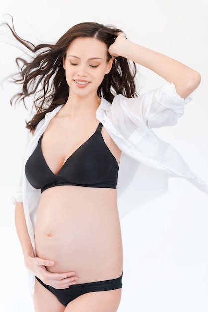 Fresh and beautiful pregnant woman. Beautiful pregnant woman holding hand in hair and touching her belly while standing against white background