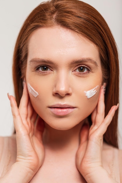 Fresh beautiful young girl with red hair applies cream on her face skin and looks at the camera Beauty and skin care