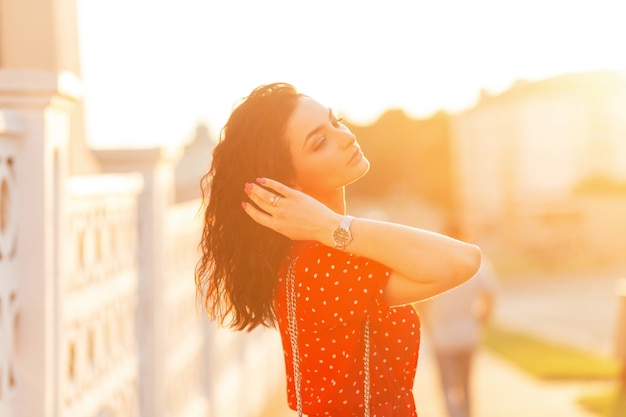 Fresh beautiful young woman in a fashionable red dress walks in the city at sunset