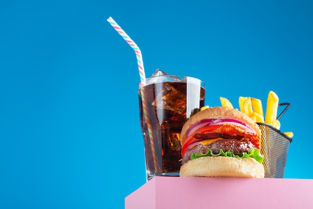 Fresh beef hamburger, cola and fried french fries placed on the pink stand and blue background. Copy space for text, trendy hero view. Horizontal orientation