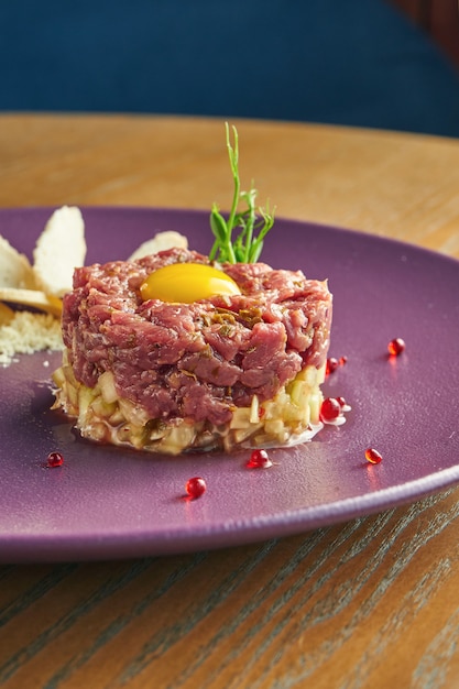 Foto tartare di manzo fresca con parmigiano, capperi, crostini e quaglia di tuorlo su un piatto viola su una parete di legno