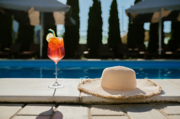 Fresh beverage in glass and hat at the edge of the pool, nobody. carefree summer vacation concept, holiday party at the poolside outdoors