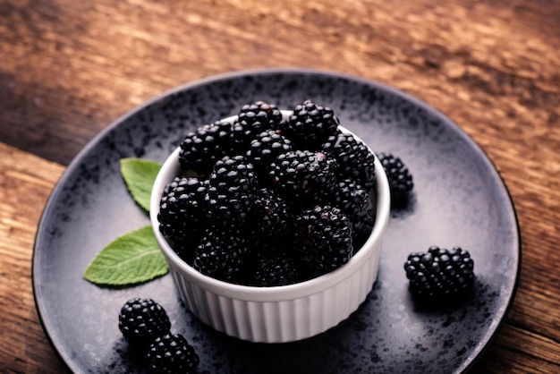 Fresh blackberries in a white bowl on a black plate on a wooden background closeup