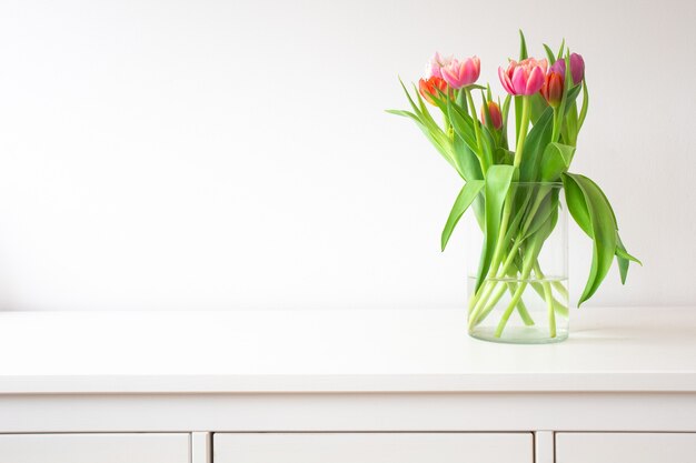 Fresh bright bouquet of colorful Tulips near white wall on wooden shelf in glass vase wit space for text