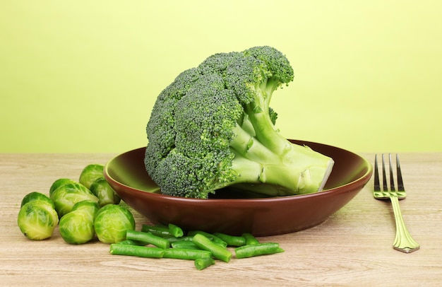 Fresh broccoli on plate with cabbages and french bean on wooden table on green background