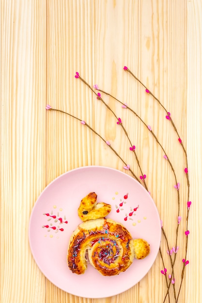 Fresh bun in the form of an Easter bunny. The concept of children's holiday bakery. In pink ceramic plate with artificial willow branches on wooden surface, copy space, top view, close up.