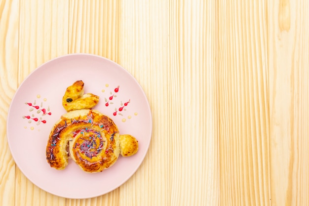 Fresh bun in the form of an Easter bunny. The concept of children's holiday food. In pink ceramic plate on wooden surface, copy space, top view.