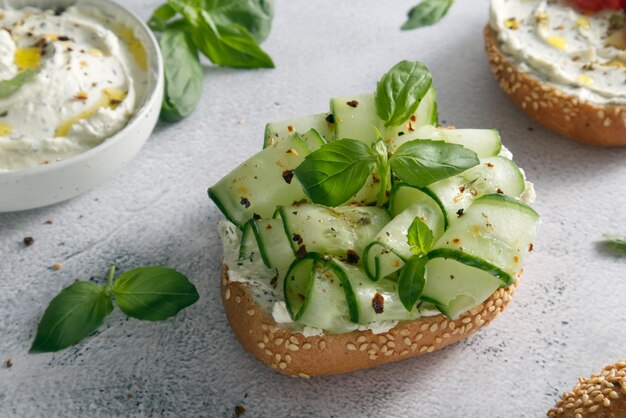 Fresh bun with sesame seeds with cheese cream and fresh cucumbers with basil leaves close-up