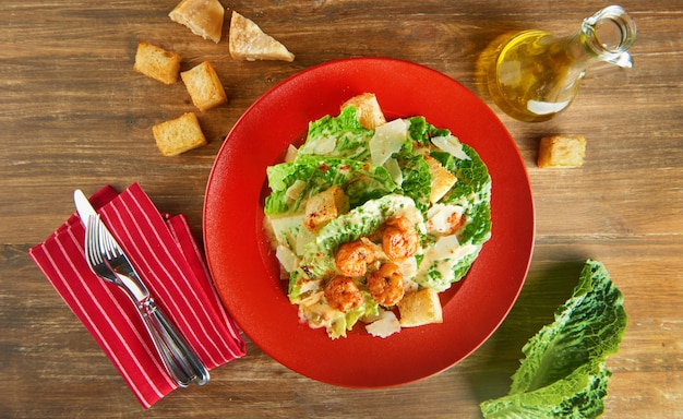 Fresh caesar salad on red plate with parmesan cheese and shrimps on wooden table. Top view