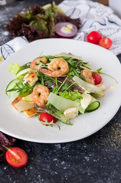 Fresh caesar salad with shrimps on a white plate on dark stone background