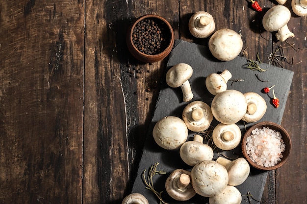 Funghi champignon freschi con spezie ed erbe aromatiche. ingrediente crudo per cucinare cibi vegani. vecchio fondo di legno, luce dura di modo, ombra scura, stile rustico, vista dall'alto