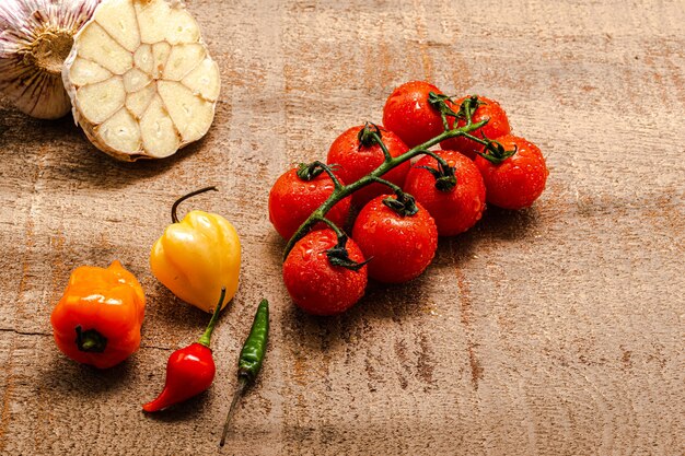 Fresh cherry tomatoes on a rustic brown wooden table with several peppers and sliced garlic around them. Top view